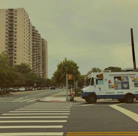 A no walk sign is lit at a crosswalk in The Bronx. Next to the sign is an ice cream truck. No people are around. To the left is a high rise apartment block.
