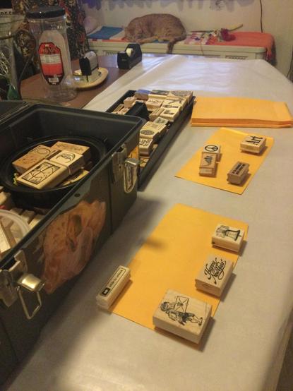 long view of table w rubber stamps arranged on envelopes to show where they’ll be printed, at top of frame orange cat sleeps on radiator, to the left you can see a large plastic toolbox w lift out tray both are fulll of rubber stamps