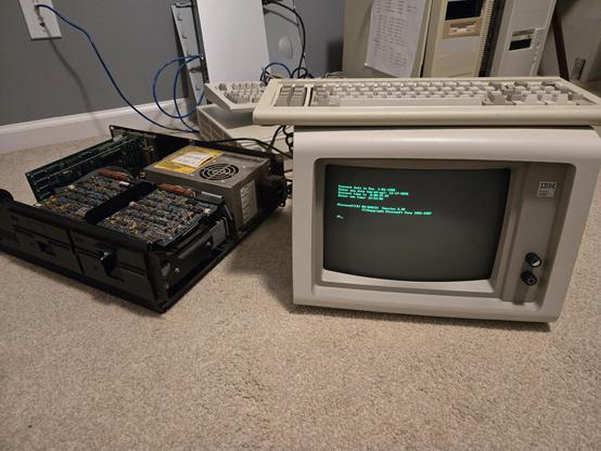 an IBM 5150 with the case open is on the left. A 5151 monitor sits on the carpet to the right. the text in the screen is the MS-DOS welcome banner from my DOS 3.3 master.
