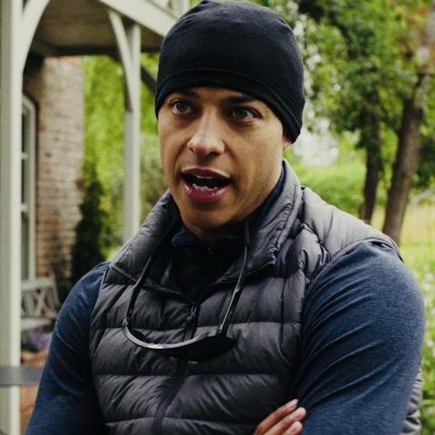 Movie still: A man (Daryl McCormack) wearing a black beanie and a grey vest stands with his arms crossed, appearing to speak or express an emotion. The background features greenery and a structure typical of a garden or outdoor setting.