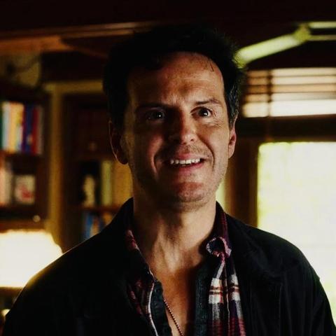 Movie still: A man (Andrew Scott) with dark hair and a slight smile, wearing a plaid shirt under a dark jacket, stands inside a cosy room filled with books and warm lighting.
