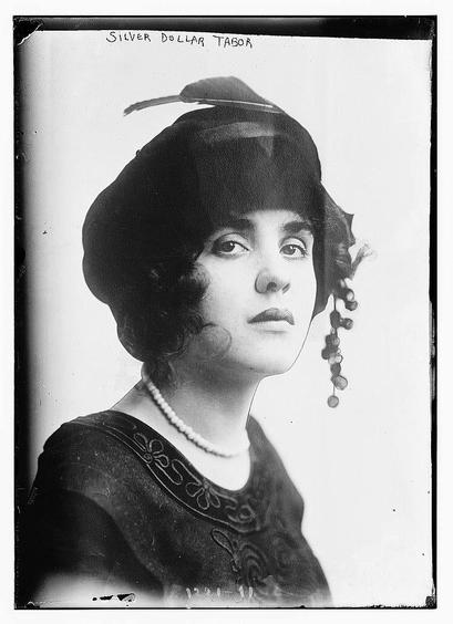 The image is a black-and-white portrait of a woman. She is wearing a dark, possibly black, hat adorned with a feather on the side. Her hair is styled in a manner typical of the early 20th century, with curls framing her face. She is also wearing a pearl necklace and a dark dress with a patterned design on the upper part. The photograph appears to be a formal portrait, likely taken in a studio setting, given the plain background and the subject's direct gaze. The text "SILVER DOLLAR TABOR" is written at the top of the image, which might be a caption or a reference to the subject or the photograph. The overall style and attire suggest that the photo was taken in the early 1900s.