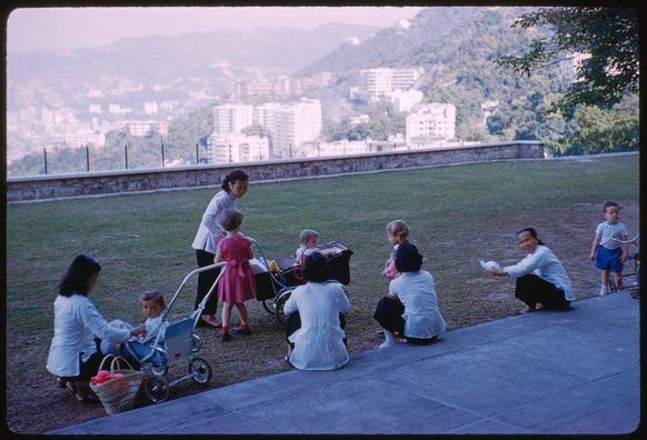 an image of a group of people outside having fun together. they all are wearing white and sitting around on the grass by some trees, with a city in back drop . this photo was taken in hong kong , january 1859 as part of sports illustrated magazine collection. it is said to be a sight seeing trip through the beautiful scenery