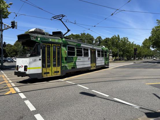 A small white and green two bogie, three (or six) door tram rolls through the street of Melbourne on a sunny day, the greenery of the botanical gardens fills the background. The tram is new enough to be metal bodied, yet old enough to have folding doors, with a typical pantograph picking up power from the overhead line. Tram tracks criss cross the road, as tracks go to and from various areas toward the city centre.