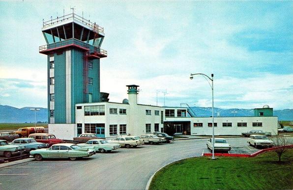 This color postcard from the mid-1960s shows Missoula County Airport in western Montana. The scene highlights the airport’s tall, narrow control tower clad in vertical green and white panels, topped with a glass-walled cab and surrounded by a red safety railing. Attached to the tower is the low, horizontally oriented terminal building, a midcentury structure with flat roofs, rectangular windows, and exterior stairways. A variety of early-1960s automobiles fill the parking lot in the foreground, including sedans, wagons, and a Shell fuel truck. Behind the airport buildings, open fields stretch toward a backdrop of mountains beneath a lightly clouded sky. The image reflects a period of expanding regional air service and modest postwar airport development in Montana.