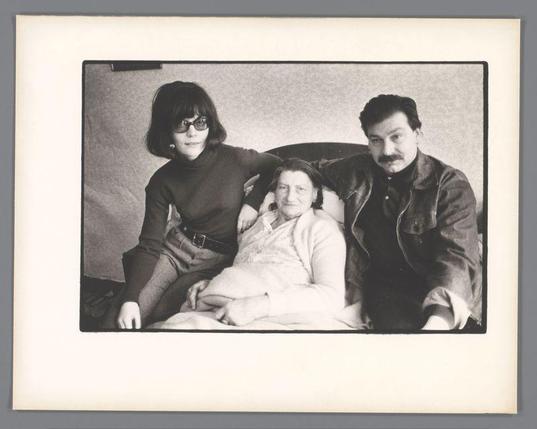 The image is a black and white photograph featuring three individuals seated closely together on what appears to be a couch. On the left, there's an individual with long hair wearing glasses, dressed in a dark sweater over a light-colored top, paired with dark pants or a skirt; their hands are clasped on their lap. The person sitting in the middle is lying down and seems relaxed, enveloped by what looks like bedding material, possibly indicating they might be unwell as suggested by one of the descriptive tags mentioning an elderly woman in bed. On the right sits another individual with dark hair styled neatly, wearing a leather jacket over layers of clothing; their expression appears contemplative or serious.

The background is plain and nondescript, focusing attention on the subjects. The photograph seems to convey a moment shared among family members during what could be considered a vulnerable time for one person involved due to health circumstances. There are no discernible objects aside from furniture that provides context about where this might have been taken, such as at home or in another personal space.

The image is framed within the boundaries of a white sheet with a visible border and clip on top left corner, indicating it's likely part of an album or collection of photographs. The overall quality suggests it could be from around mid-20th century based on clot [...]