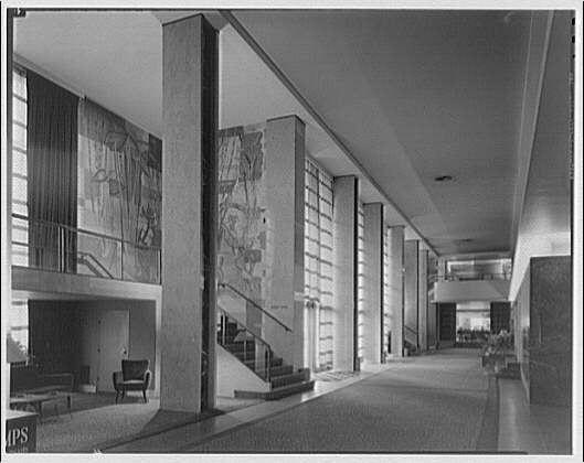 The image depicts the interior of a grand building, likely an early to mid-20th-century structure given its architectural style. The main focus is on two massive vertical columns with abstract paintings adorning their sides, adding artistic flair and character to the space. These columns are part of a larger open-plan area that extends into the distance, featuring wide walkways lined by benches for seating.

Natural light floods in through large windows or possibly glass doors at the end of the hall, indicating an entrance or exit point beyond view. The floor appears polished with reflective qualities, enhancing the overall brightness and spaciousness of the lobby. There are a few people visible in the distance, suggesting that it is not overly crowded but has some activity.

A staircase on one side leads up to another level, hinting at multiple floors within this establishment. On the left-hand side near the bottom corner, there's an inscription "IPS," which may be related to the photograph or a marking specific to the building itself. The overall ambiance is that of elegance and simplicity, with clean lines dominating the design aesthetic.

The black-and-white nature of the photo implies it could be historical documentation rather than contemporary imagery.