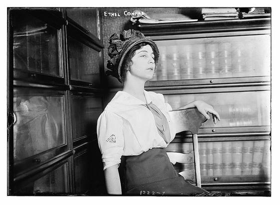 The image is a black-and-white photograph of a woman seated in a chair in what appears to be a library or study setting. She is wearing a white blouse with puffed sleeves and a dark apron. Her attire suggests a professional or academic environment, possibly related to research or study. She is also wearing a decorative hat with a large bow on top, which adds a touch of elegance to her appearance. The background features a bookshelf filled with numerous books, reinforcing the academic or scholarly context. The woman is looking slightly to her right, and her expression is calm and composed. The photograph has a vintage quality, and there is text at the top of the image that reads "ETHEL CONRAD," which is likely the name of the person depicted. The overall tone of the image is formal and suggests a historical setting.