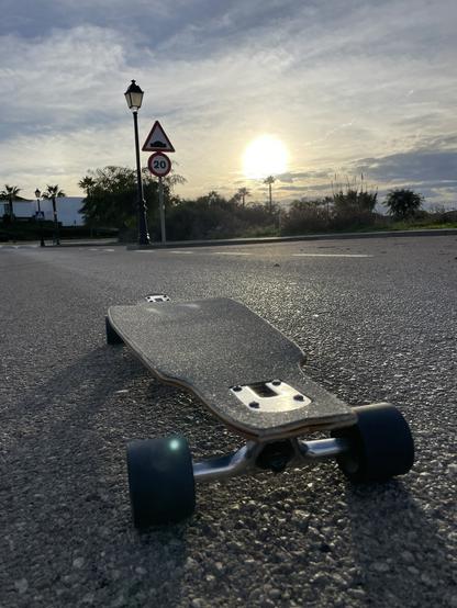Das Longboard auf Höhe des Boards von hinten gegen die tief stehende Sonne fotografiert. Im Hintergrund säumen Büsche und Bäume die Straße.