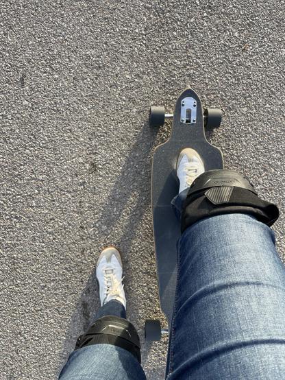 Blick von oben aufs Board. Mein rechter Fuß steht darauf, der linke auf dem Boden. Ich trage weiße Turnschuhe, Bluejeans und schwarze Knieschoner.