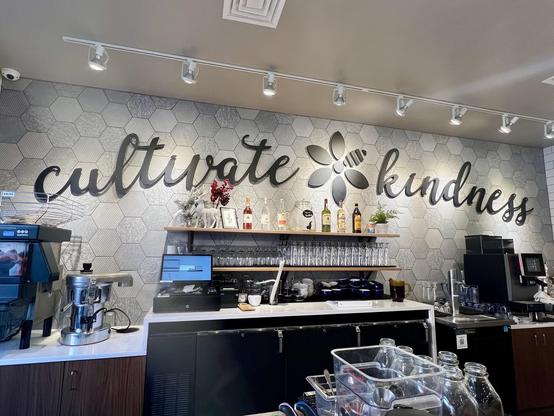 A coffee shop interior featuring a wall with the phrase "cultivate kindness" in elegant lettering. Below this, there is a wooden shelf displaying various bottles and glasses. Equipment for making drinks is visible, including a coffee machine and a blender.