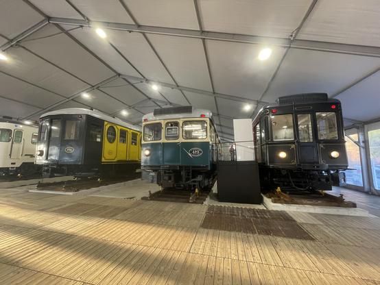 Three historical trains are displayed inside a large tent. The trains include a yellow and black one in the left, a teal and white train in the centre, and a black train on the right. The flooring is wooden, and the space is well lit.