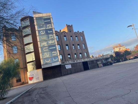 An angled view of a cultural center “La Farinera” featuring modern and historical architectural elements. The building has large windows and prominent signage. The surrounding area includes a paved space and landscaping, with a clear sky above.