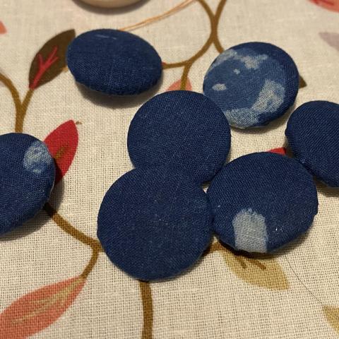 Buttons covered in navy blue fabric with paler blue markings one of which looks like a face. They are on a patterned vinyl tablecloth.