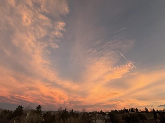 A wide-angle photo of sweeping clouds near sunset. Clouds are in pink, orange, salmon colors, and there's one airplane contrail in the same color.