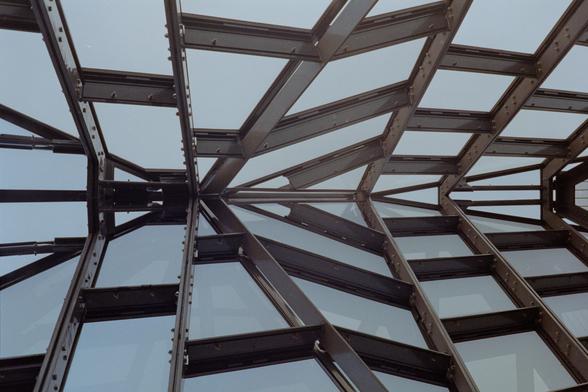A color photograph of abstract geometric shapes which upon closer inspection reveal they're metal trusses holding up glass window panes at some interesting angles. The perspective is rotated from vertical to horizontal but the shapes still don't quite make sense as a triangular roof with panels. The photo is cooler on the left side and warmer on the right.
 Shot with a Canon CanonetG-IIIQL17 and Canon40f1.7 lens, on FujiC200, and developed with BelliniC41 by Shom Bandopadhaya. Licensed under Creative Commons Attribution-NonCommercial-ShareAlike (CC BY-NC-SA).