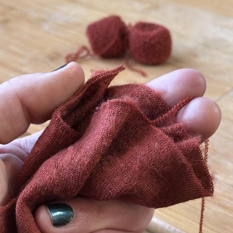 Eine Hand mit dunkelgrün glitzernd lackierten Nägeln hält den halb aufgeribbelten Ärmel eines rost-orangen Merino-Feinstrickpullovers in die Kamera.

Von der aufgeribbelten Kante kräuselt sich ein hauchfeiner Wollfaden über die Hand nach unten. 

Im verschwommenen Hintergrund liegen zwei kleine Knäuel des wieder aufgewickelten Garns.