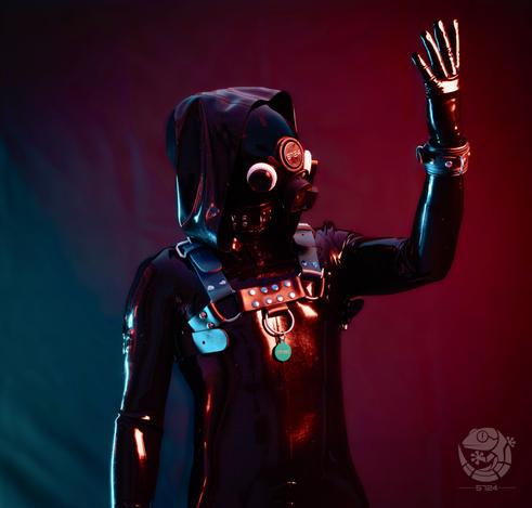 A black latex drone entity stands against a red and blue lit background, wearing a full latex suit with hood, gas mask and padded harness. The surface of the suit reflects the colored lights like polished synthetic material. One gloved arm is raised with the hand open, as if signaling or acknowledging a command. A metal ring and tag hang from the chest harness. The face is fully obscured, removing any human identity and presenting the subject as a controlled, functional unit in a club environment.