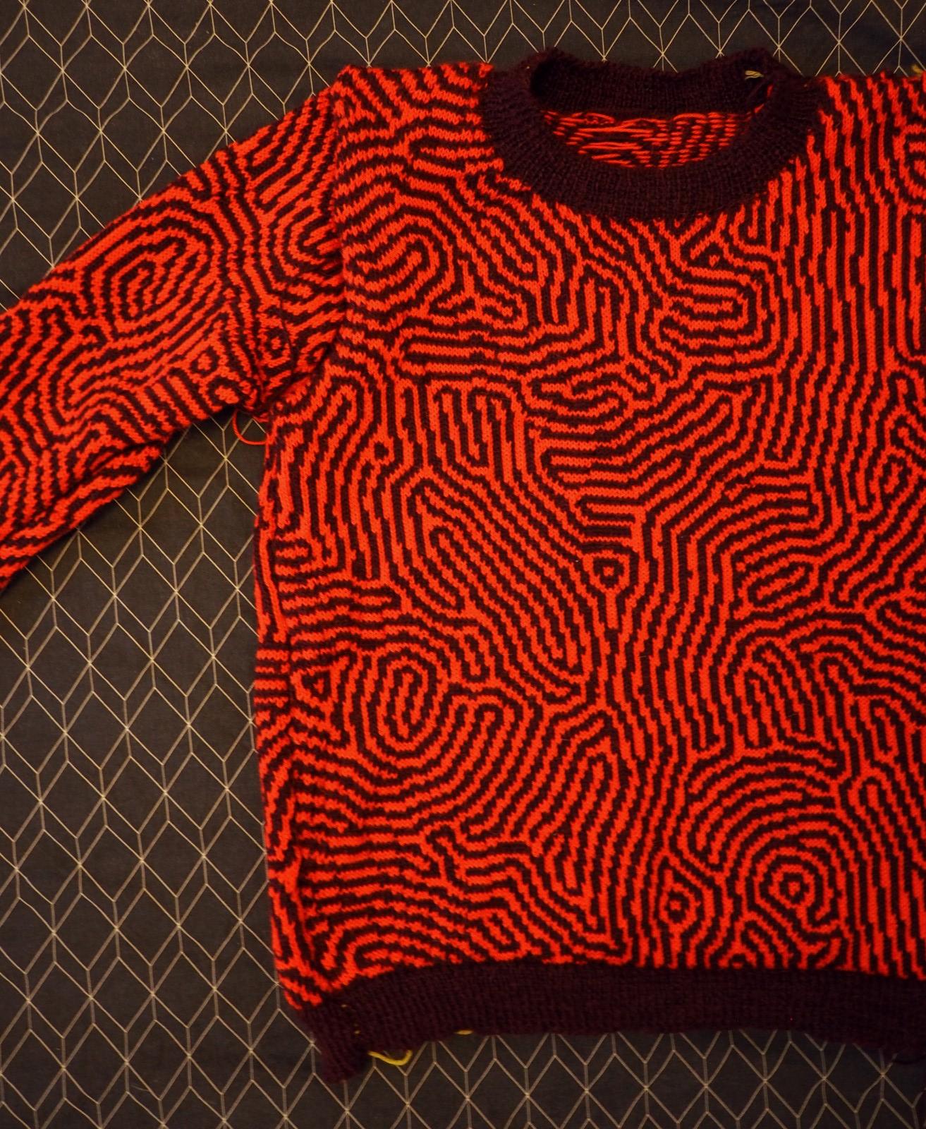 A knitted sweater laid flat and viewed from above. The design on the knit is a bright red diffusion reaction pattern on a dark red background.