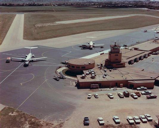 This is an aerial photograph of the Lubbock Municipal Airport taken in the late 1960s or early 1970s. The image looks down on the terminal, control tower, aircraft ramp, and surrounding airfield. The terminal is a long, low tan building with a flat roof. Near the center is a round, two-story section with curved windows. Behind it stands a square tan control tower with small rectangular windows and an open deck. Above the deck is a glass-walled cab with radar antennas and communication masts on the roof.

Two commercial jets are parked on the ramp. Both are narrow-body jets with rear-mounted engines, likely early DC-9s. One jet is positioned close to the curved terminal section with ground service equipment nearby. The other is parked farther back, near the edge of the ramp. A small red-and-white airport vehicle is driving near the aircraft in the foreground.

The ramp markings are visible: wide taxiway lines, hold-short lines, and parking stand outlines. Beyond the ramp are long concrete runways and taxiways surrounded by dry, brownish grass. In the lower part of the image is a small parking lot filled with cars and pickup trucks of late 1960s styles. The horizon shows the city of Lubbock in the distance with low buildings and flat terrain.