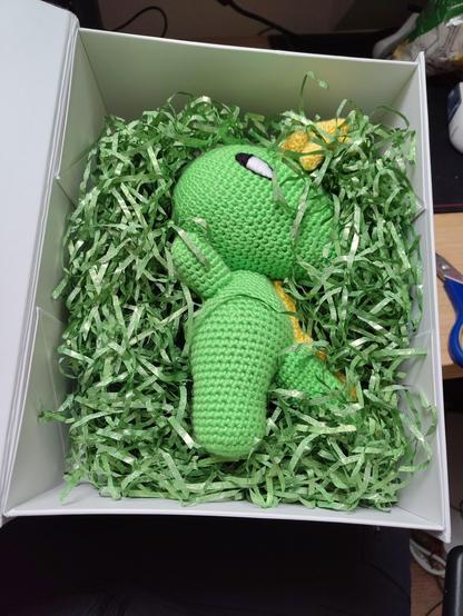 the box has been opened, it shows green packing fill, surrounding a beautifully hand knitted green Konqi plushie
