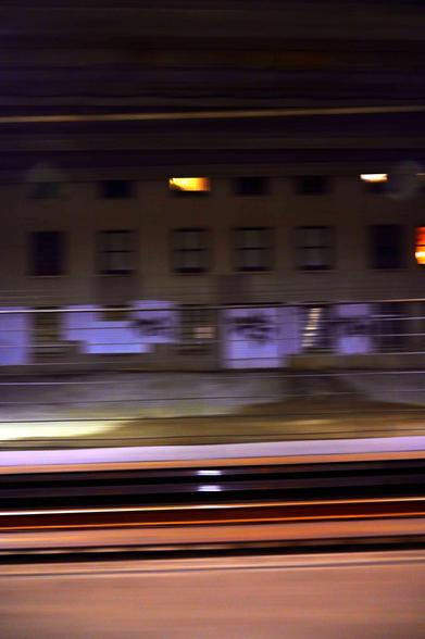 Train tracks — two power rails, two running rails, and an induction strip — rushing by from the perspective of another train. in the background, a blurry building can be seen, with blue lights along its ground floor and windows above it. The whole space looks like it's underground.