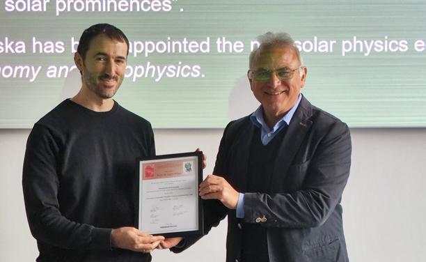 Two people present a framed certificate in front of a screen displaying text.