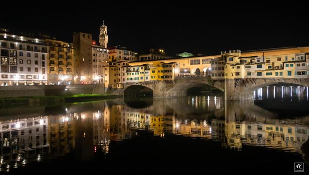 Nighttime color photo of an old bridge covered with buildings over the Arno River in Florence. The bridge and the buildings along the far side are reflected in the still waters of the river.