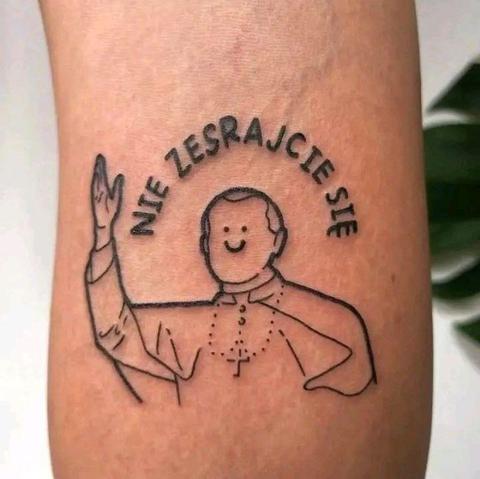 Na zdjęciu widać fragment ludzkiego ramienia z prostym tatuażem w czarnym kolorze.

Na środku jest bardzo schematycznie narysowana postać duchownego w ornacie, z uniesioną prawą ręką w geście błogosławieństwa. Twarz jest uproszczona: okrągła głowa, dwie małe kropki zamiast oczu i półokrągły uśmiech, bez innych szczegółów. Na piersi widać zarys różańca zakończonego krzyżykiem.

Nad głową postaci, po łuku, znajdują się duże drukowane litery tworzące napis po polsku: „NIE ZESRAJCIE SIĘ”. Napis i rysunek są cienką, równą linią, bez cieniowania czy koloru wypełniającego.