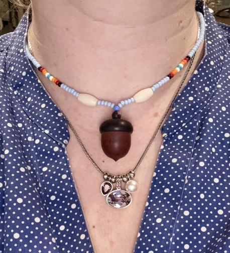 A close-up of my chest area with two necklaces. The top necklace is a beaded necklace with a wooden acorn pendant. The beads are light blue with accents of red and white. The lower necklace is a silver chain with several little charms. One charm is a large, horizontal, oval, pink gem. Another charm is a little pearl. And the last charm is another pink gem but smaller.