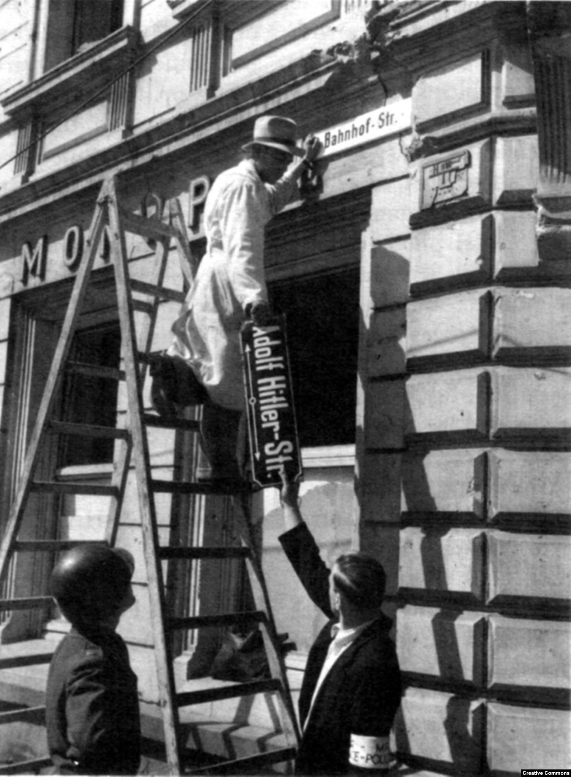 Man on a ladder, wearing a hat and a white trenchcoat, passing down a sign that says “Adolf Hitler Str.”