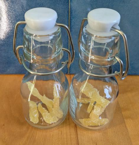 The image displays two small swing top glass bottles with pieces of crystallized ginger inside sitting on top of a blonde waxed cutting board with blue handmade ceramic tile in the background.