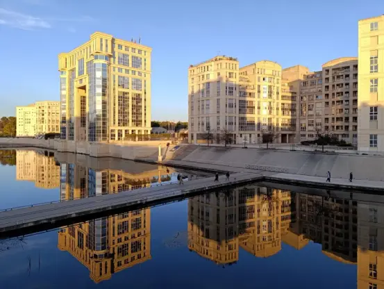 Photo prise des hauteurs d'un pont au-dessus d'un petit fleuve dont l'eau est canalisée par des bassins. La lumière du soleil couchant se déverse sur des immeubles du quartier Antigone de Montpellier. Architecture librement inspirée de celle de la Grèce antique. Le ciel est bleu. On distingue quelques personnes qui marchent sur la passerelle et sur les rives du Lez.