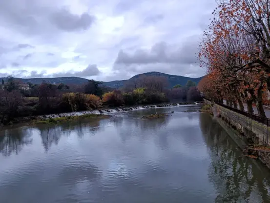 Un cours d'eau avec une rangée de platanes sur la droite de l'image. Une petite cascade artificielle traverse le fleuve dans sa largeur. Au fond, un petit massif sous un ciel très nuageux.