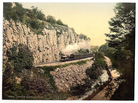The image depicts a vintage scene featuring a steam train in motion, traveling through a narrow valley. The locomotive is emitting smoke as it climbs along the tracks that wind alongside rocky cliffs on one side and greenery and trees on another. A small stream or waterway meanders parallel to the railway track below. The photograph has an aged quality with sepia tones typical of old postcards, suggesting its historical nature from around 1890-1900. Notable features include the natural landscape surrounding the train tracks and the rustic charm evoked by the steam locomotive's presence in a rural setting.