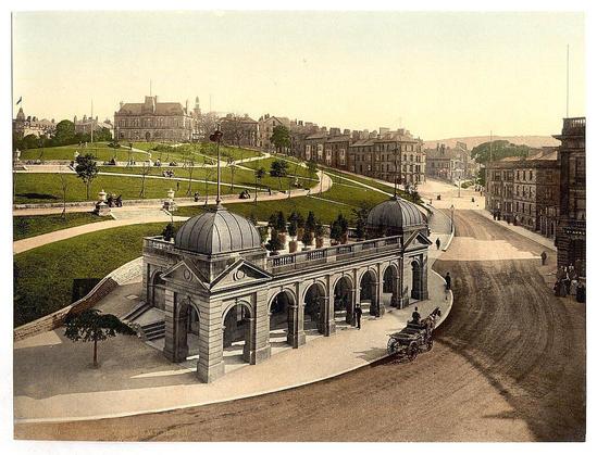 The image depicts a historical scene from Buxton, specifically focusing on the pump room in Derbyshire, England. It appears to be an old postcard or photochrome print dating between approximately 1890 and around 1900. The photograph captures a well-maintained public square with classical architecture prominently featured.

In the center of the image is an ornate building resembling a small domed pavilion, likely serving as a pump room for its historical context. It features several arches under a semi-circular colonnade and multiple windows that allow light to filter through, creating a serene atmosphere.

The surrounding area includes neatly manicured lawns with pathways winding up the gentle slopes of what seems like park grounds or terraces. People can be seen walking on these paths, dressed in period clothing suggestive of late 19th-century fashion. A horse-drawn carriage is present at the bottom right corner, adding to the historical ambiance.

The background reveals a town-like setting with rows of buildings and distant structures that could be residential housing or institutional architecture such as schools or government offices. The scene conveys an urban environment from another era, providing insight into how public spaces were designed in Buxton during this time period.