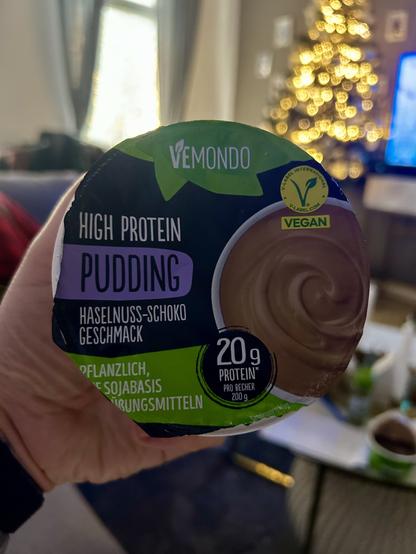 Auf dem Bild hält eine Hand einen Vemondo High Protein Pudding in der Geschmacksrichtung Haselnuss-Schoko. Der Pudding ist vegan, auf Sojabasis und enthält 20 g Protein pro 200-g-Becher. Der Deckel ist überwiegend dunkel gehalten mit grünen und violetten Akzenten sowie einem Bild des cremigen Schokopuddings.

Im Hintergrund ist ein wohnlicher Innenraum zu sehen: ein beleuchteter Weihnachtsbaum mit warmem, unscharfem Lichterbokeh, ein Fenster mit Vorhängen und ein Fernseher auf der rechten Seite. Die Umgebung wirkt gemütlich und leicht weihnachtlich, während der Fokus klar auf dem Puddingbecher im Vordergrund liegt.