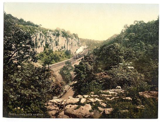 The image depicts a scenic landscape with lush greenery and rocky terrain. In the foreground, there is an area filled with large rocks and boulders scattered across uneven ground, suggesting a rugged outdoor setting. Beyond this rocky expanse lies a dense thicket of trees and shrubs, presenting various shades of dark greens and browns.

Dominating the middle portion of the scene is a railway track cutting through the landscape. The tracks are laid out in such a way that they follow the natural topography, curving around hillsides as it extends into the distance. A steam train is captured in motion on this rail line; its presence adds a dynamic element to an otherwise static environment.

The backdrop features towering cliffs or rocky crags adorned with vegetation, indicating a remote and possibly challenging terrain for railway construction. The sky above has a hazy quality, suggesting either early morning light or the onset of dusk, which casts a muted glow over the entire scene.

At the bottom left corner, there is text that reads "10863 - OHEEDALE, BUXTON," indicating the specific location as Buxton's Oheedale. The combination of natural beauty and human engineering in this setting provides an evocative snapshot of life during a period when railway expansion was transforming landscapes across Britain.

This image is part of a collection known for its vivid photographic [...]