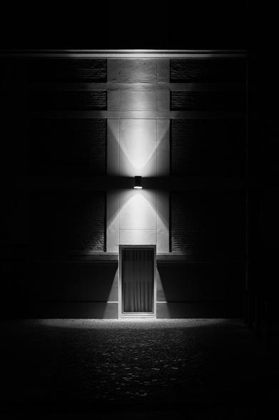 A dimly lit wall with a door at the center, highlighted by a single overhead light. The scene is captured in black and white, creating a dramatic, shadowy atmosphere.