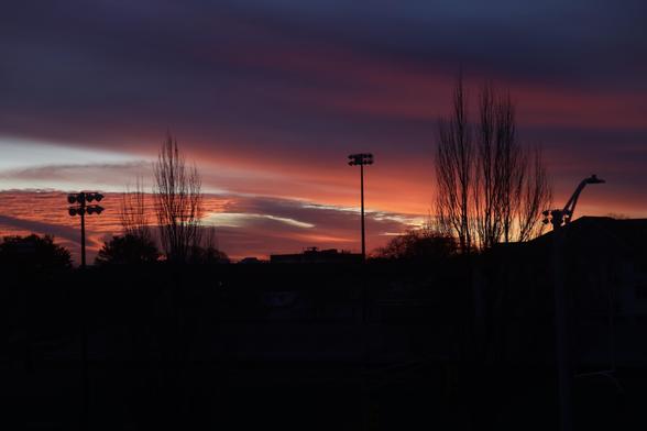 A vibrant sunset with shades of orange, pink, and purple fills the sky, silhouetting trees and stadium lights against a dark foreground.