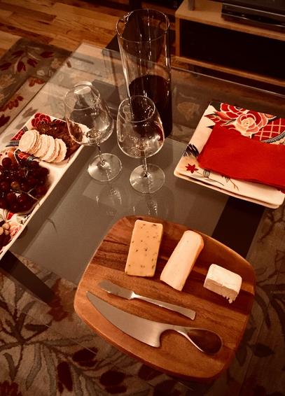 A small square glass table has hors devours set up comprised of cheese and crackers, grapes, and red wine. There are two wine glasses, two square plates and red napkins. The cheese is on a wooden cutting board with a couple of knives. The lighting is low.