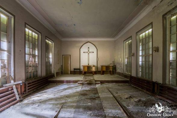 An abandoned chapel shows the decay of time. Light floods in through large windows, illuminating the space with a cross above the altar.