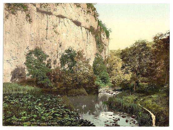 The image depicts a tranquil rural landscape with lush greenery. A prominent, weathered cliffside rises in the background on one side of the scene, while trees and bushes dot another part leading into the distance. In the foreground, there's an idyllic river reflecting the surrounding foliage, bordered by dense vegetation including what looks like aquatic plants near its edge. The water meanders gently through a rocky stream bed with stones protruding above it. A curved dirt path or trail is visible on the right-hand side of the image leading into the distance. Overall, this scenic view appears to be an early 20th-century photograph capturing the serene beauty of Derbyshire's countryside.
