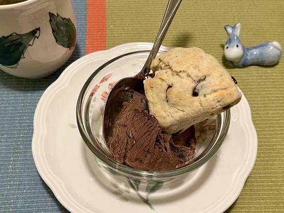 A small glass bowl containing a scoop of chocolate ice cream topped with mini scone sits on a white plate. Nearby, there is a ceramic cup and a small figurine of a bunny. They’re on the placemat.