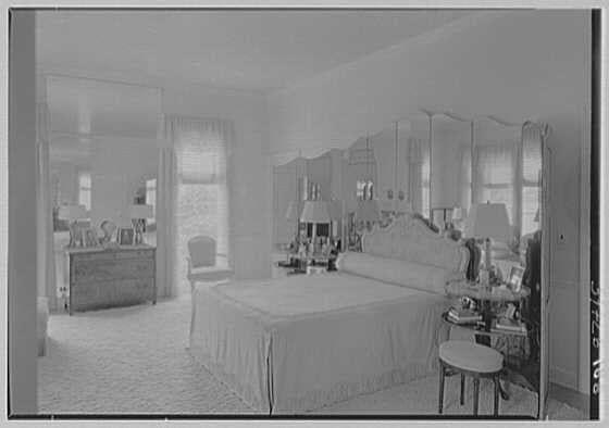 The image depicts a vintage bedroom setting, as evidenced by the black and white photograph. The room appears to be well-furnished with a large bed positioned against one wall, covered in what looks like light-colored bedding or drapes. To the right of the bed is an ornate vanity table with a mirror above it; this piece seems to have several items on top, including lamps and possibly cosmetics.
The window behind the bed allows natural light to illuminate the room through sheer curtains that hang from rod supports near the ceiling. A chair can be seen in front of one side of the large dresser or chest located against another wall opposite the bed. The flooring is carpeted, adding a touch of coziness to the space.
A stool with no backrest stands next to the vanity table on its right side while an armchair sits beside it. In the background, there's another chair by the window and various other items scattered throughout the room such as books or magazines, which suggest that this might be a personal living area where someone spends time relaxing.
Overall, the bedroom exudes a classic elegance with tasteful decor reflecting its historical context.