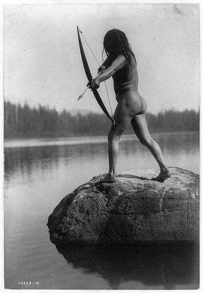The image is a black-and-white photograph showing an individual, likely from the Nootka culture in British Columbia during the early 19th century. The person stands on a rock in calm waters and is drawn back with a bow, aiming it as if preparing to shoot an arrow into the distance. They wear minimal clothing—a brown sleeveless garment that reveals their upper body—and have long hair tied up away from their face. Their legs are barefooted, emphasizing the simplicity of life during this period.

The background features what appears to be forested land across a broad expanse of water, suggesting a remote or wilderness setting. The photograph carries historical significance and reflects on indigenous practices before widespread European contact in North America. This particular image is part of a collection named "The Bowman—Nootka c1910," which provides further context about its cultural origin.

This photographic print measures 235 mm by 348 mm, indicating it's likely intended for display or archiving purposes rather than mass reproduction at the time. Edward S. Curtis (1868-1952) is renowned as a photographer who documented Native American cultures through his extensive work in ethno-photography during the late 19th and early 20th centuries, making this image an important piece of cultural heritage for understanding historical indigenous communities.