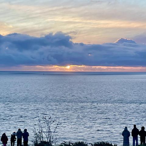 A serene sunrise over the ocean, with colourful clouds above and a few people silhouetted in the foreground watching the view.