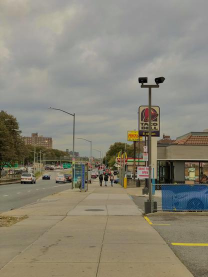 An absolutely enormous sidewalk with some students wearing backpacks in the distance walking past a Taco Bell. The sky is gloomy. A road sits to the left.