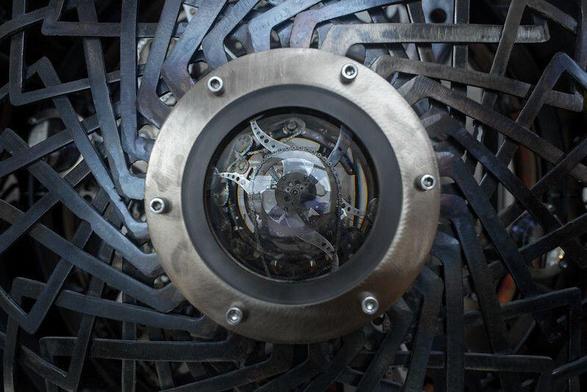 A metal ring with six rivets centered inside a complex metal latticework; inside the ring is a glass dome with complex clockwork gears and levers visible.