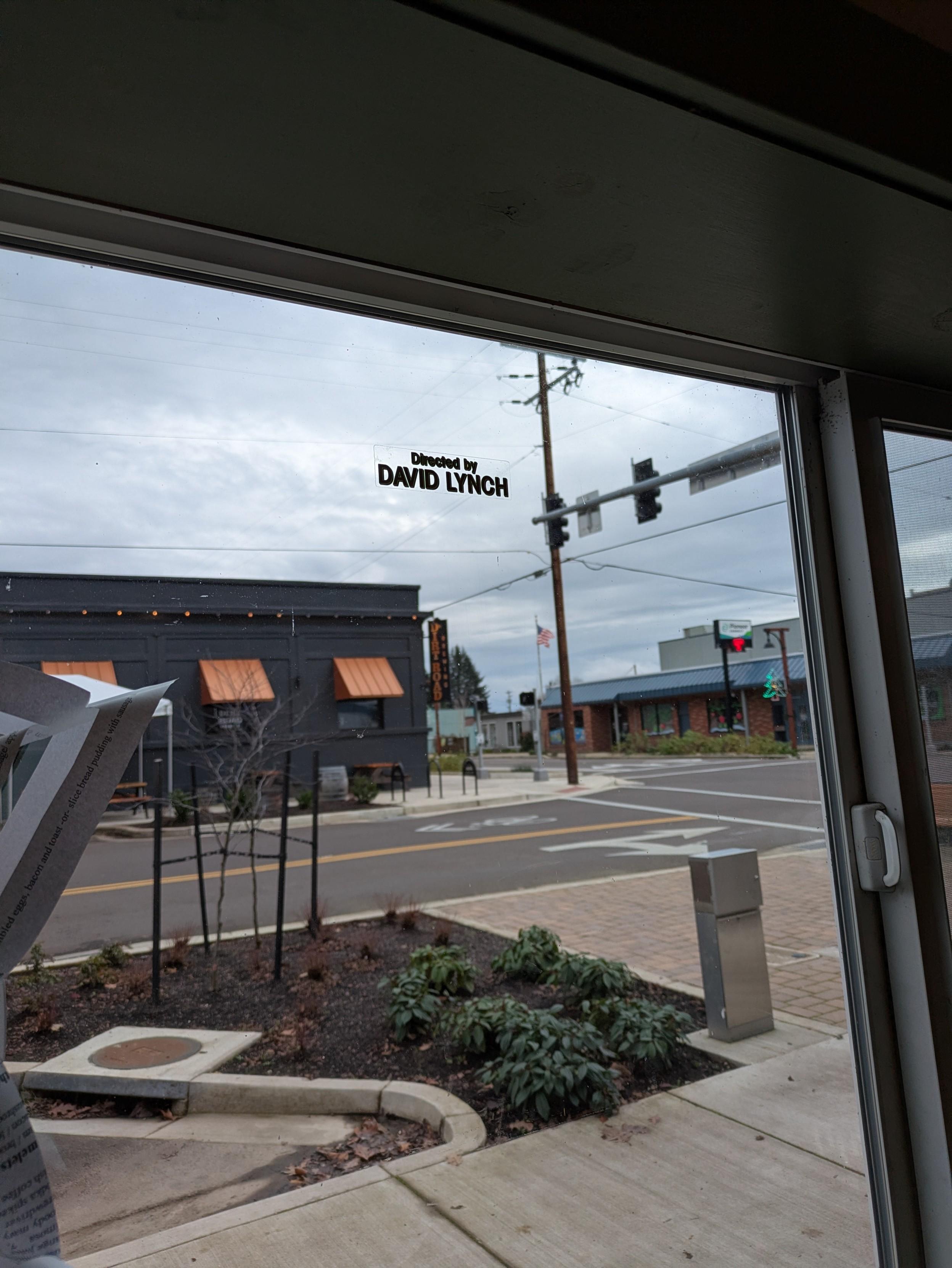 Sticker says "Directed by David Lynch" in the twin peaks font. It's placed on a window looking out over a rural Oregon town